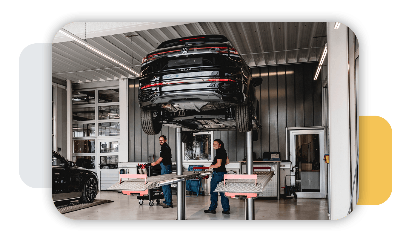 zwei Leute in der VW Werkstatt, ein Auto steht auf eine Hebbühne